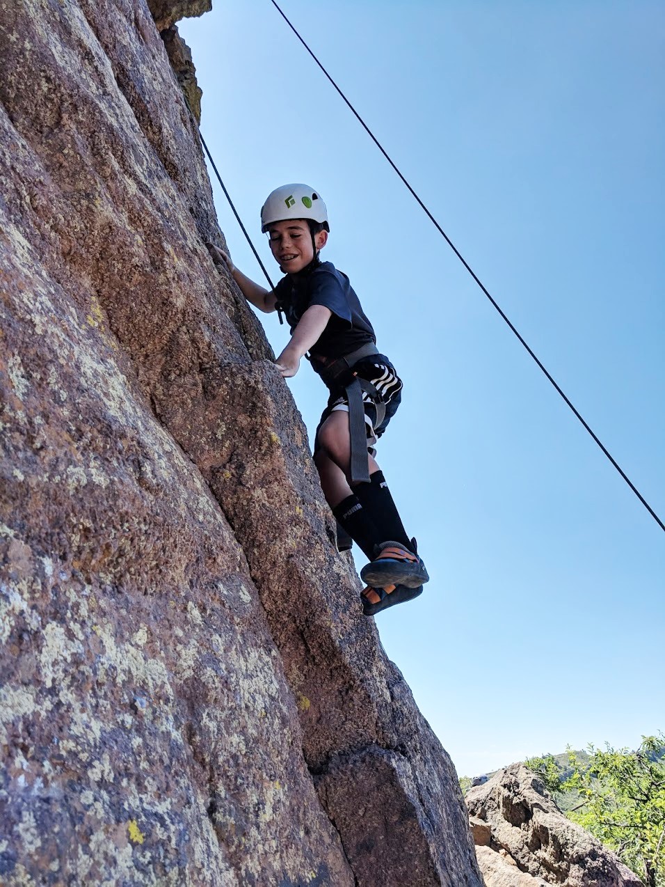 A young boy wearing a helmet and harness climbs up a cliff at Duncan's Ridge by Horse tooth Reservoir. 