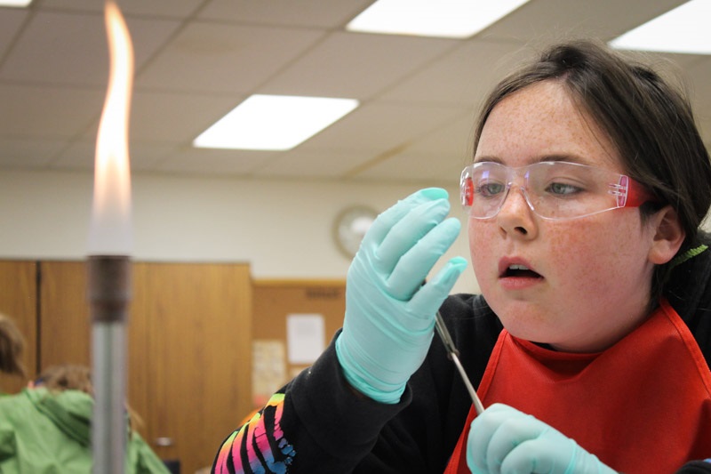 A girl with freckels wearing safety goggles examines fibers beside a Bunsen Burner.