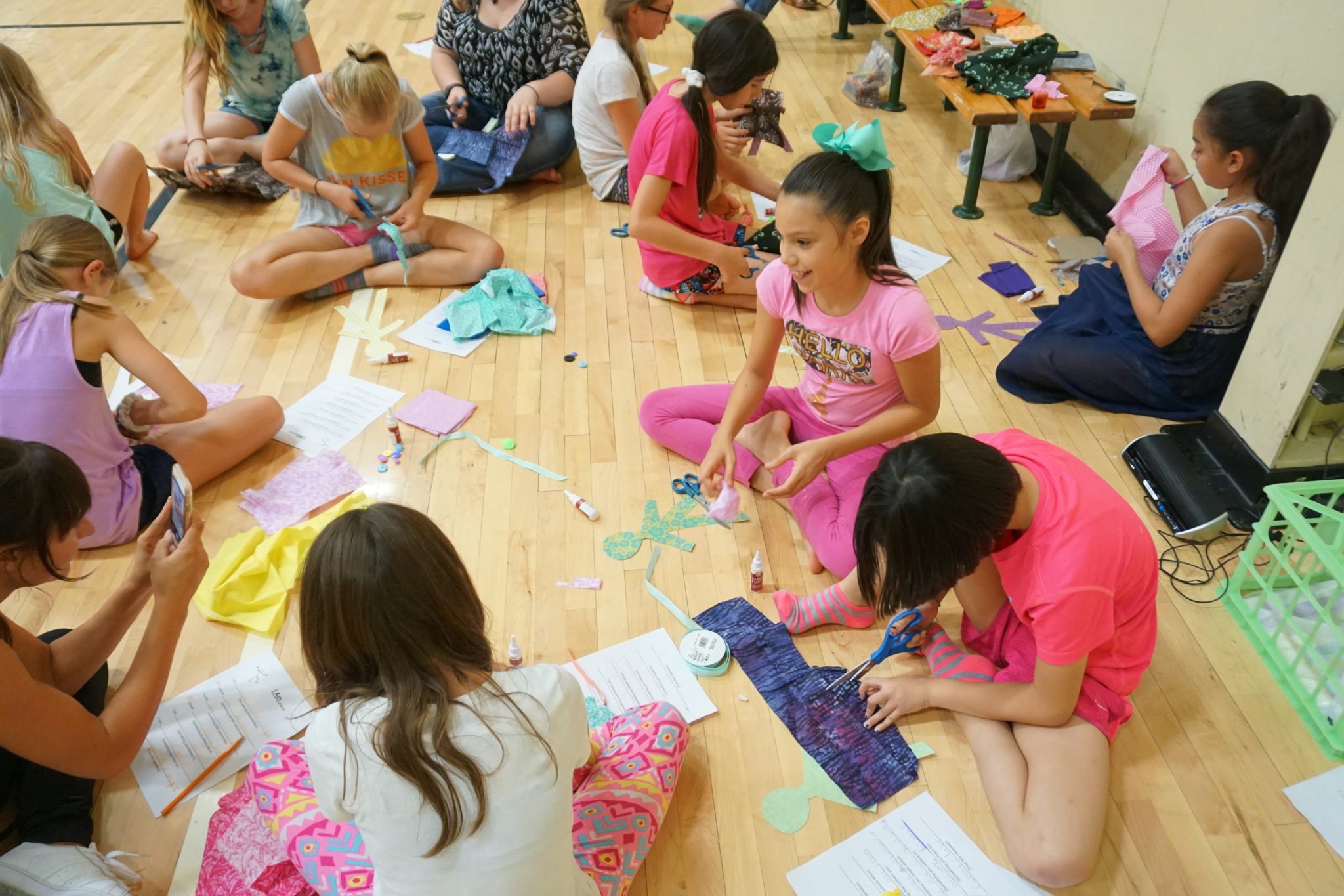 Girls wearing bright colors while seated cross-legged on a wooden floor cut fabrics and laugh as a group.
