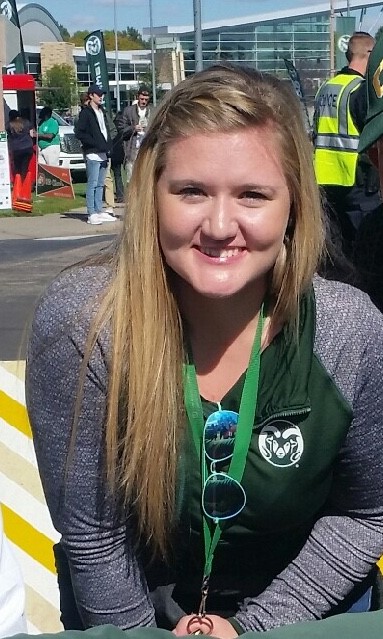 A blonde girl smiles into the sun, clad ingreen and grey rams gear and aviator sunglasses
