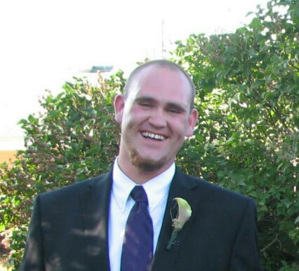 Jesse Lyall smiles as he stands in front of greenery wearing a suit.