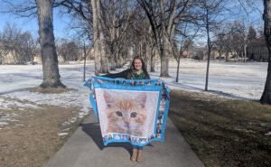 Caley stands on the Oval holding up her blanket, which features a large cat photo and a border of student mentor photographs.