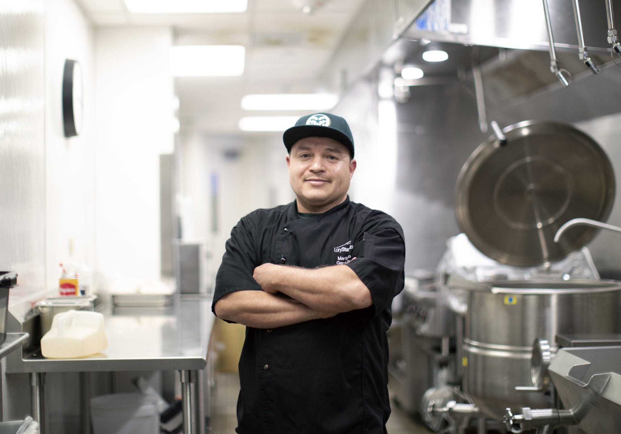 A man in a CSU hat stands with arms crossed inside an industrial kitchen.