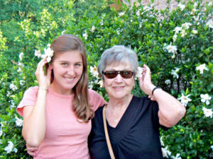 Grayson and her grandmother stand in front of flowering bushes.