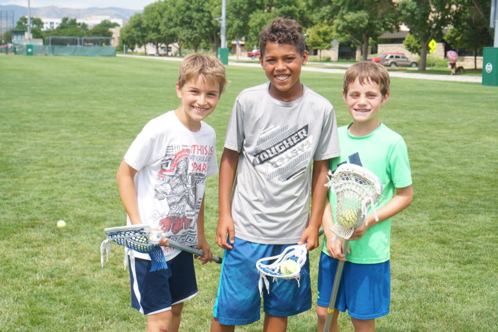 Three boys smile on a lacrosse field with lacrosse sticks.