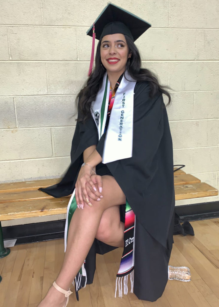 Grace Jimenez seated in the CSU auxiliary gym in her graduation regalia. 