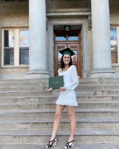 Julie Phillips, wearing a white dress and a green mortar board, stands in front of the CSU Administration Building holding her green diploma portfolio.