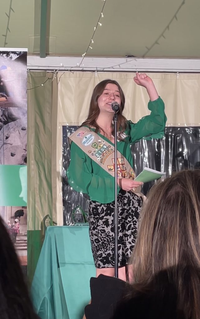 Elizabeth Gumper speaks on stage at an event. She wears a green top and a Girl Scout sash.