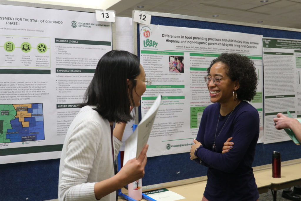 Two women stand in front of a research poster talking and laughing