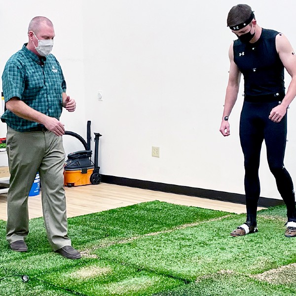 CSU professor and student walk on artificial turf indoors.