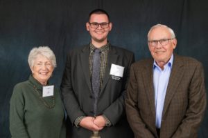 Three smiling people, posing together in front of a dark solid background: an older woman, a tall young man in glasses with a nametag that reads "scholarship recipient," and an older man in a sportcoat and glasses.