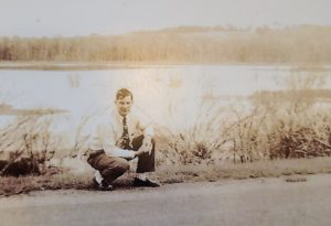 A black and white photo of a man in a tie and dress clothes crouching at the side of a road in front of a lake.