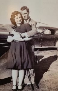 A black and white photo of a man hugging a woman with a car in the background.
