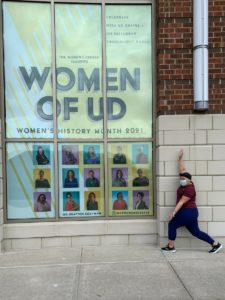 A person spreads their arms wide around a window with a large sign that reads 'Women of UD' that includes photos.