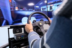 View over the left shoulder of a person holding a steering wheel in a driving simulator, with video screens showing a street at night.