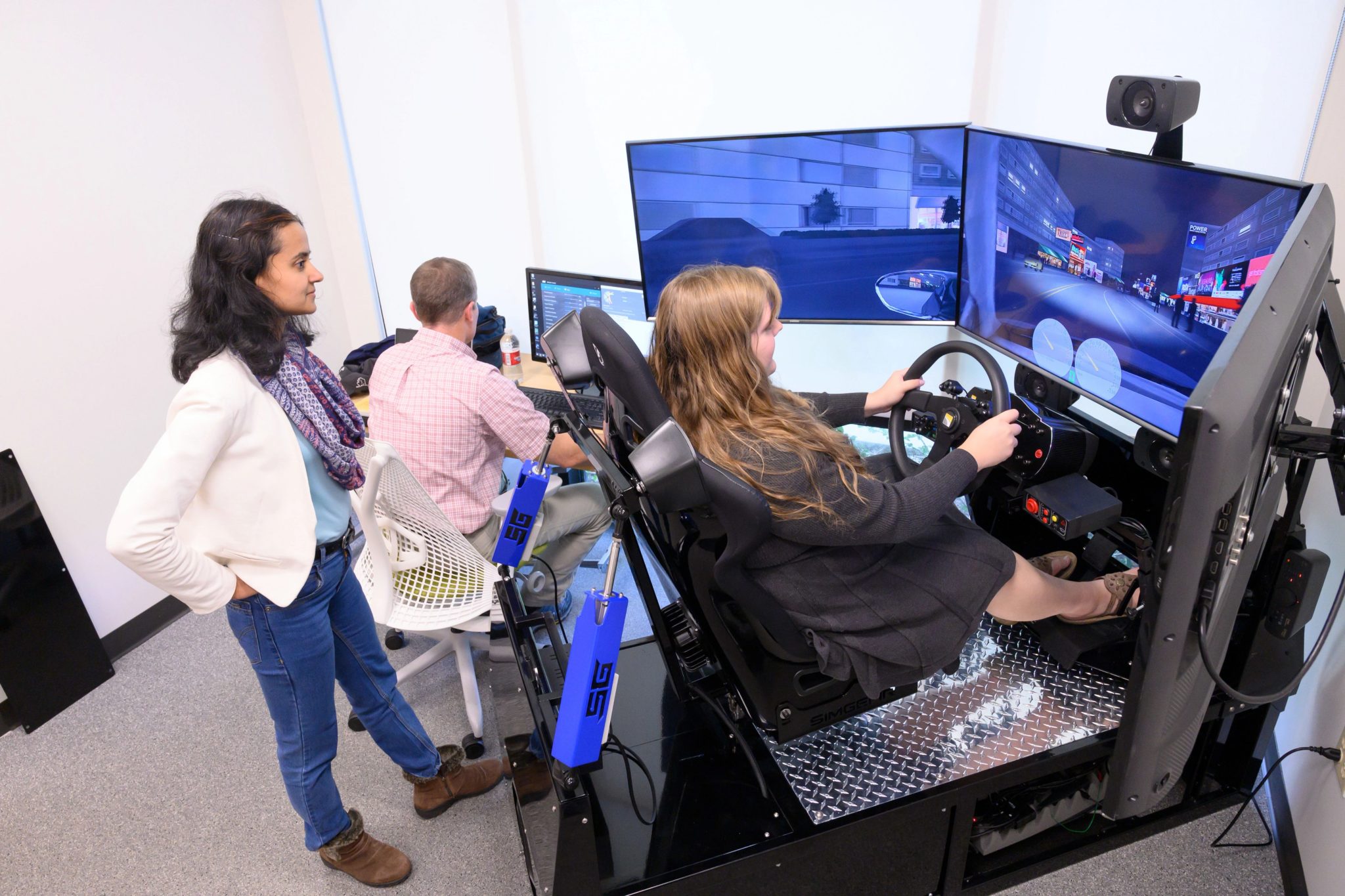 Woman stands behind another woman who is seated in a driving simulator. A man is seated at a control console to their left.
