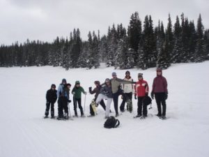 Ten people in snow gear and snowshoes stand in a field of snow in front of an evergreen forest