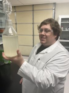 A man in a lab coat and goggles holds a clear bottle filled with cloudy liquid