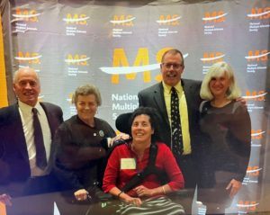 Five people - four standing, one in a wheelchair - in front of a National MS Society banner