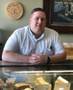A man stands behind a gourmet cheese display case.