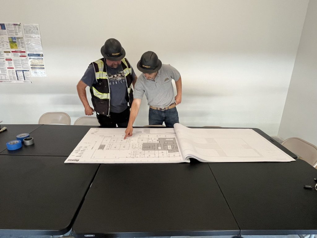 Adrian Contreras and one of his colleagues stand at a table and point at a blueprint of a construction site.