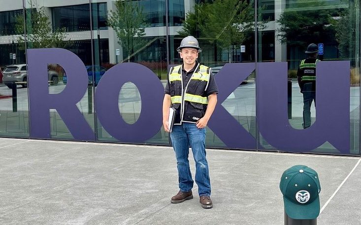 Adrian Contreras stands in construction equipment and a hard hat with the Roku building behind him.