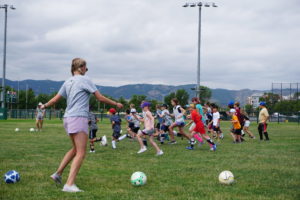 On the field, one camp counselor is kicking soccer balls toward the campers as they all run in the same direction