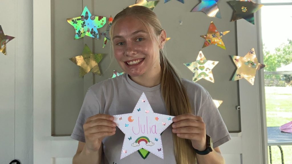 Julia Buntin is smiling standing inside of a house. She is holding up a star with her name colored on it with several other colorful stars on the wall behind her