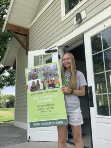 Julia Buntin is standing outside holding up a green poster with information about the FCCC Adventure camp. The posted also had four pictures on it which show kids at the camp as well as the counselors.