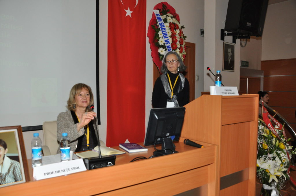 Biringen stands behind podium with Turkey flag behind her as a professor seated holds a microphone and is introducing Biringen