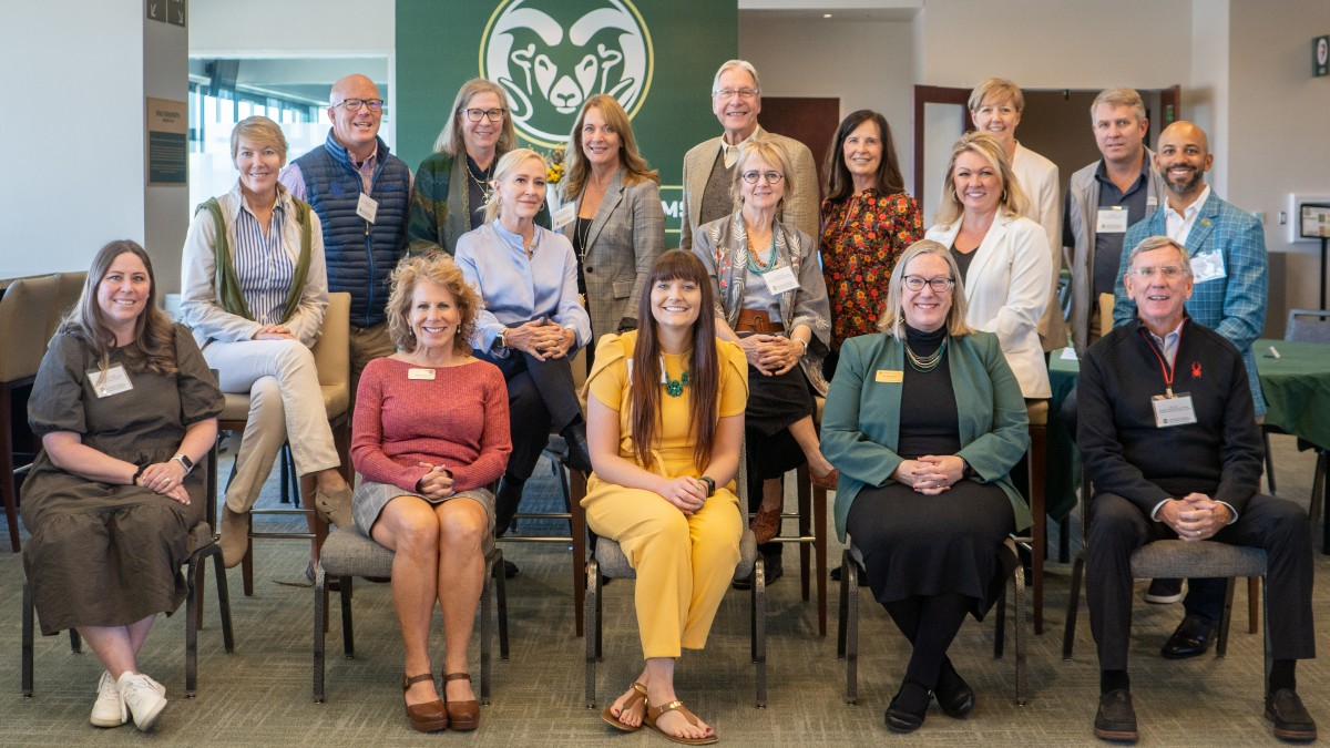 A group of adults poses, sitting and standing, for a group photo in front of a green wall with a CSU Ram's Head logo in white.
