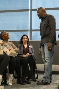 A seated Black woman looks up and smiles at a standing black man while a white woman looks on