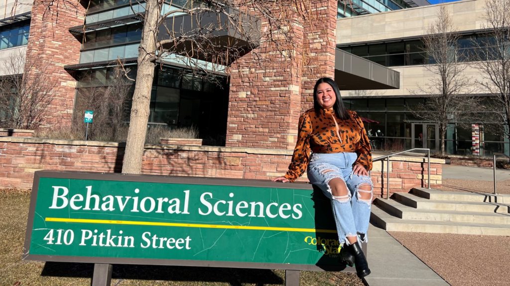 Ana Lucia Samayoa Ramos sits on green sign that reads "Behavioral Sciences, 410 Pitkin Street" in front of the Behavioral Sciences Building.