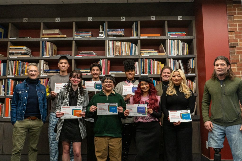 Student award winners and judges pose for a picture in the Avenir Resource Library holding awards in front of shelves of books