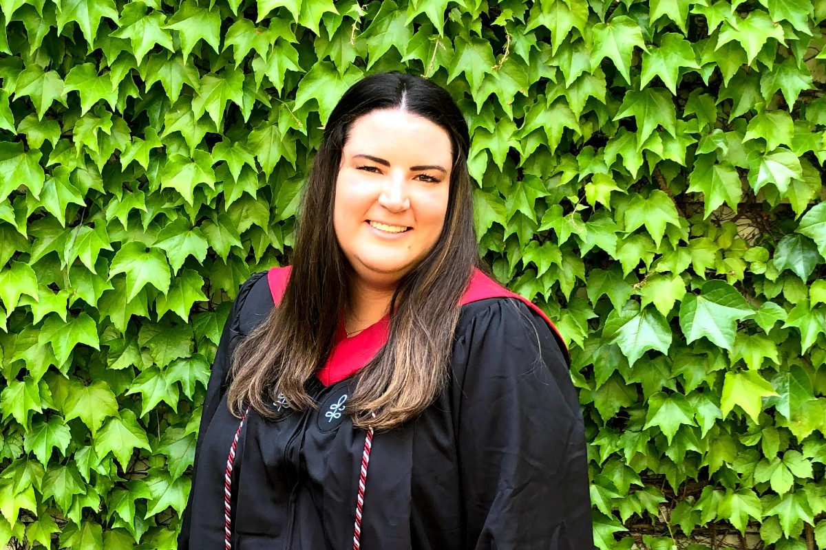 A woman with long brown hair wearing master's degree graduation regalia stands in front of an ivy-covered wall.