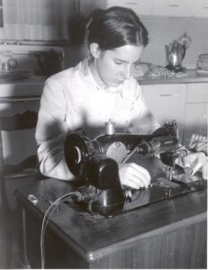 A black-and-white photo of a 15-year-old girl sitting at a sewing machine in a kitchen.