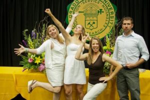 Three women in silly poses at a graduation ceremony while a man stands by