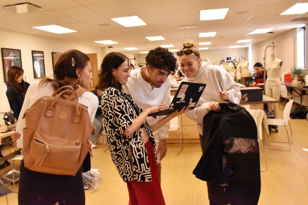 students look at a computer while looking through clothes at a model fitting
