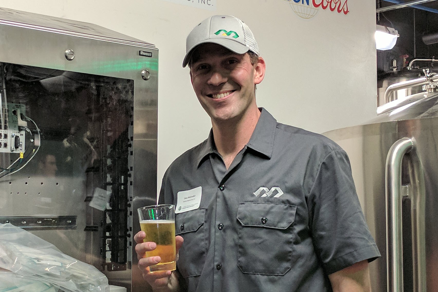 A man in a ballcap and shirt with Malisko Engineering branding holds a pint glass of beer in a brewery