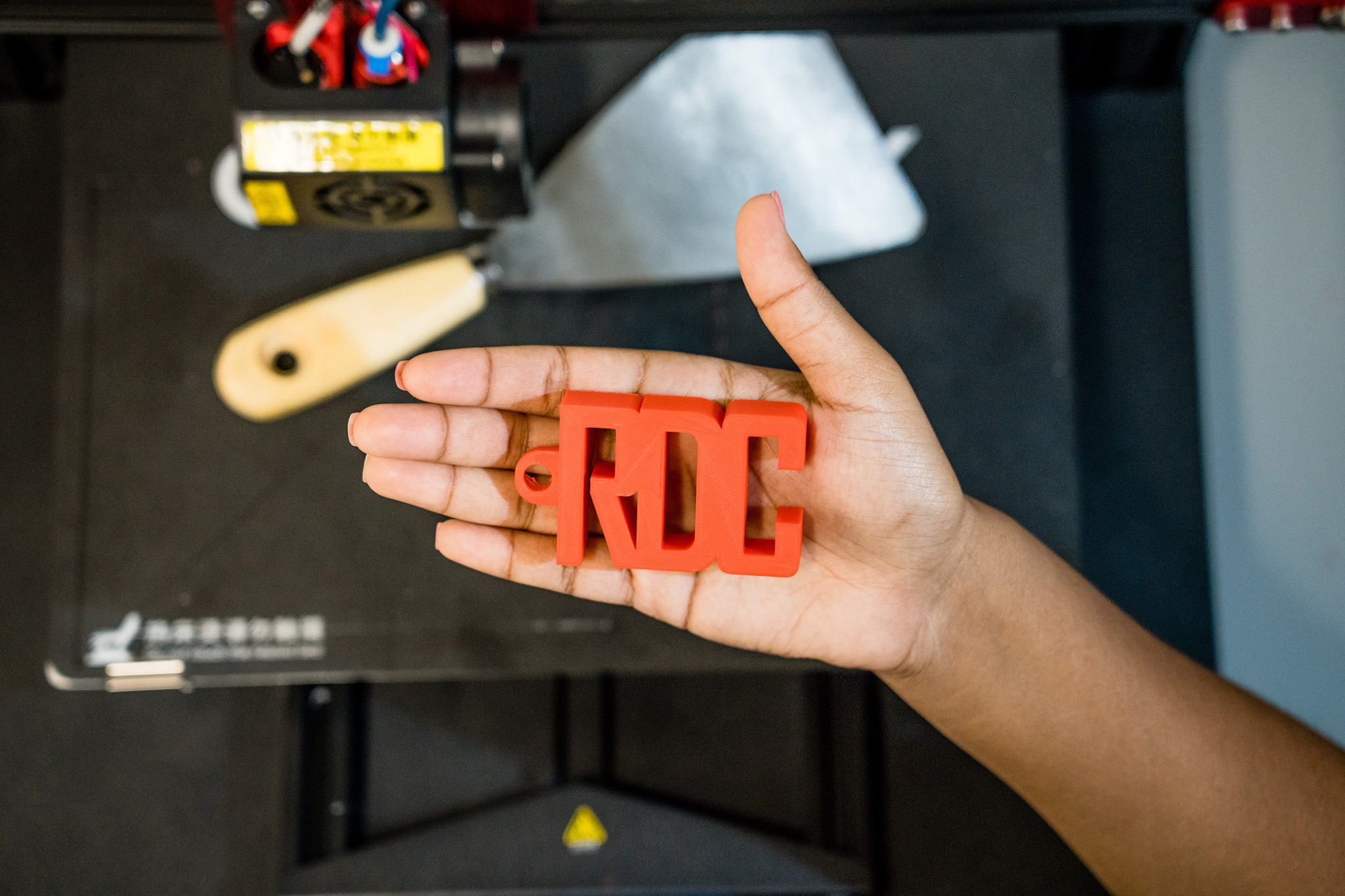 A hand holds a red red object shaped with the letters RDC, over the 3D printer and tools.