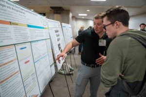 A student points to his research poster while talking with a symposium attendee