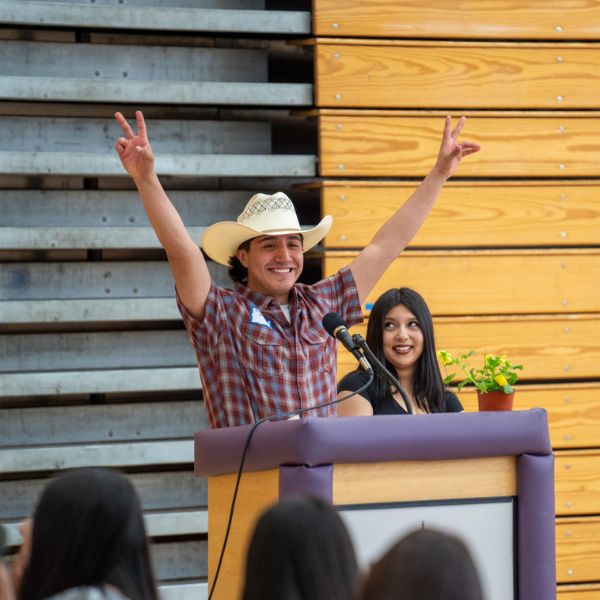 A person in a cowboy hat, standing behind a podium, with arms raised and showing the peace hand signs
