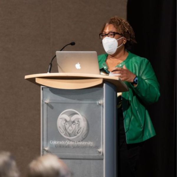 Henrika McCoy wearing a mask standing behind a podium while speaking to a group