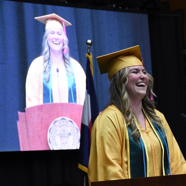 Kaylee Rookey speaking at commencement wearing cap and gown, with herself on screen behind her