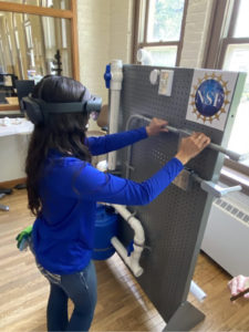 A student with virtual reality glasses looking at a mock-up wall of pipe fittings