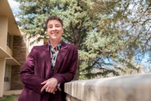 Quinn Hafen, wearing a purple suit jacket and colorful shirt, poses on a staircase in front of the Colorado State Univeristy Education building.