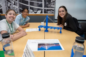 Three girls sitting at a table with their completed lego bridge