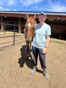 Carli stands next to a brown horse in front of a stall