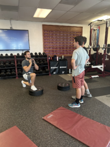 Danny does a lunge with a kettlebell in front of two high school students.