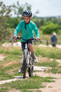 A Child in a bright teal shirt, riding a bike at a outdoor bike park.
